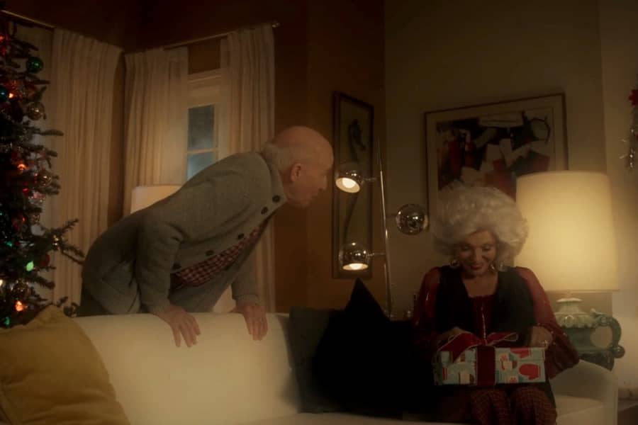 a fancy older woman with white hair opens a gift as her husband leans over the couch to watch