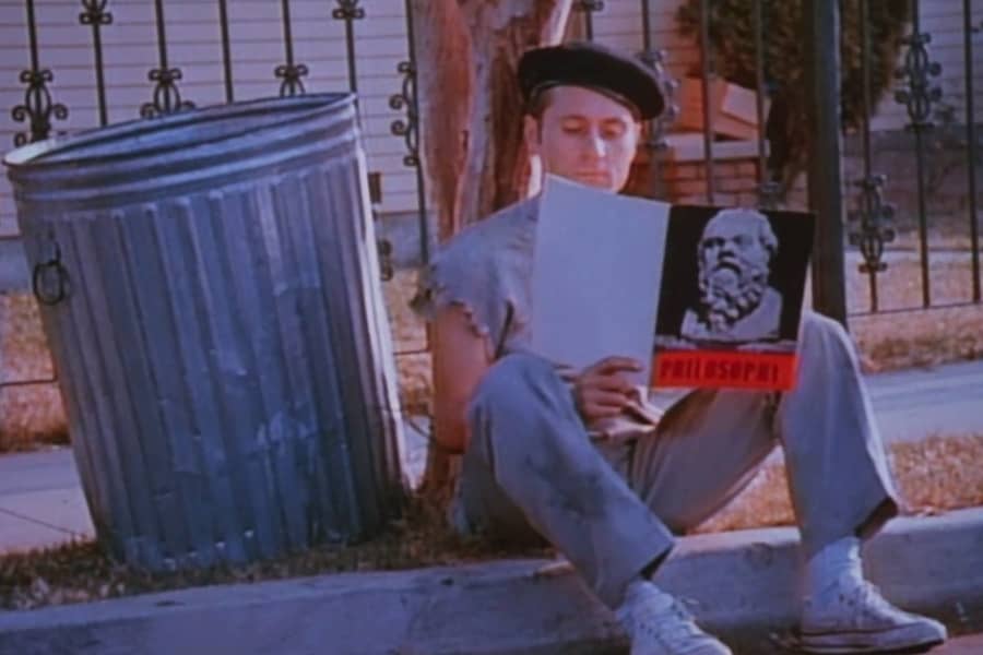 a man wearing a beret sits next to a trash can and reads a philosophy book