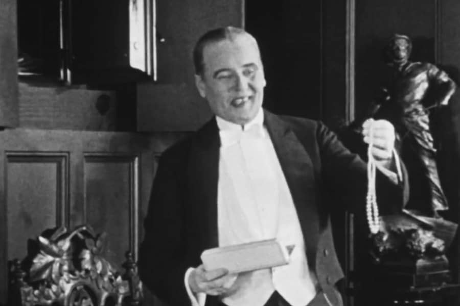 an older man holds a pearl necklace near a wall safe