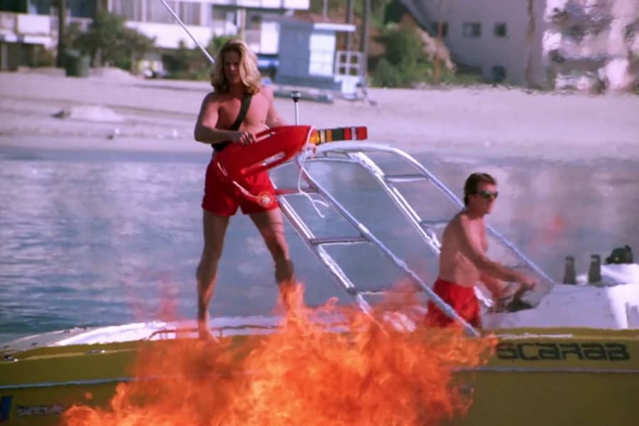 a male lifeguard stands on the edge of a speed boat as a fire burns in the foreground