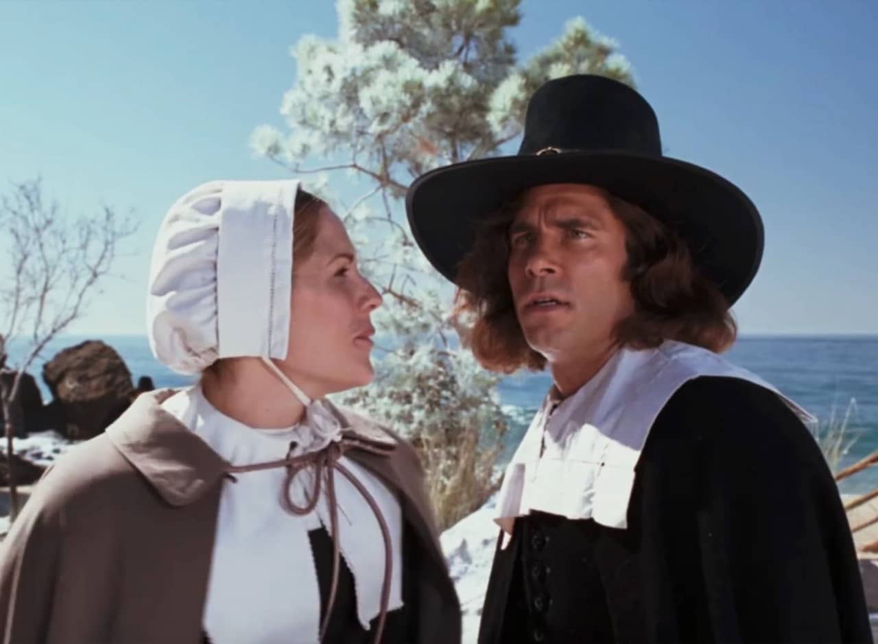 two Pilgrims, a man and a woman, talk with the ocean in the background